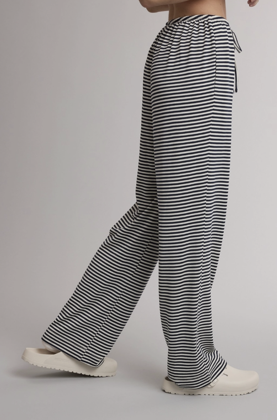 A person wearing loose black-and-white horizontally striped drawstring pants and white slip-on shoes, shown from the waist down against a plain light gray studio background.