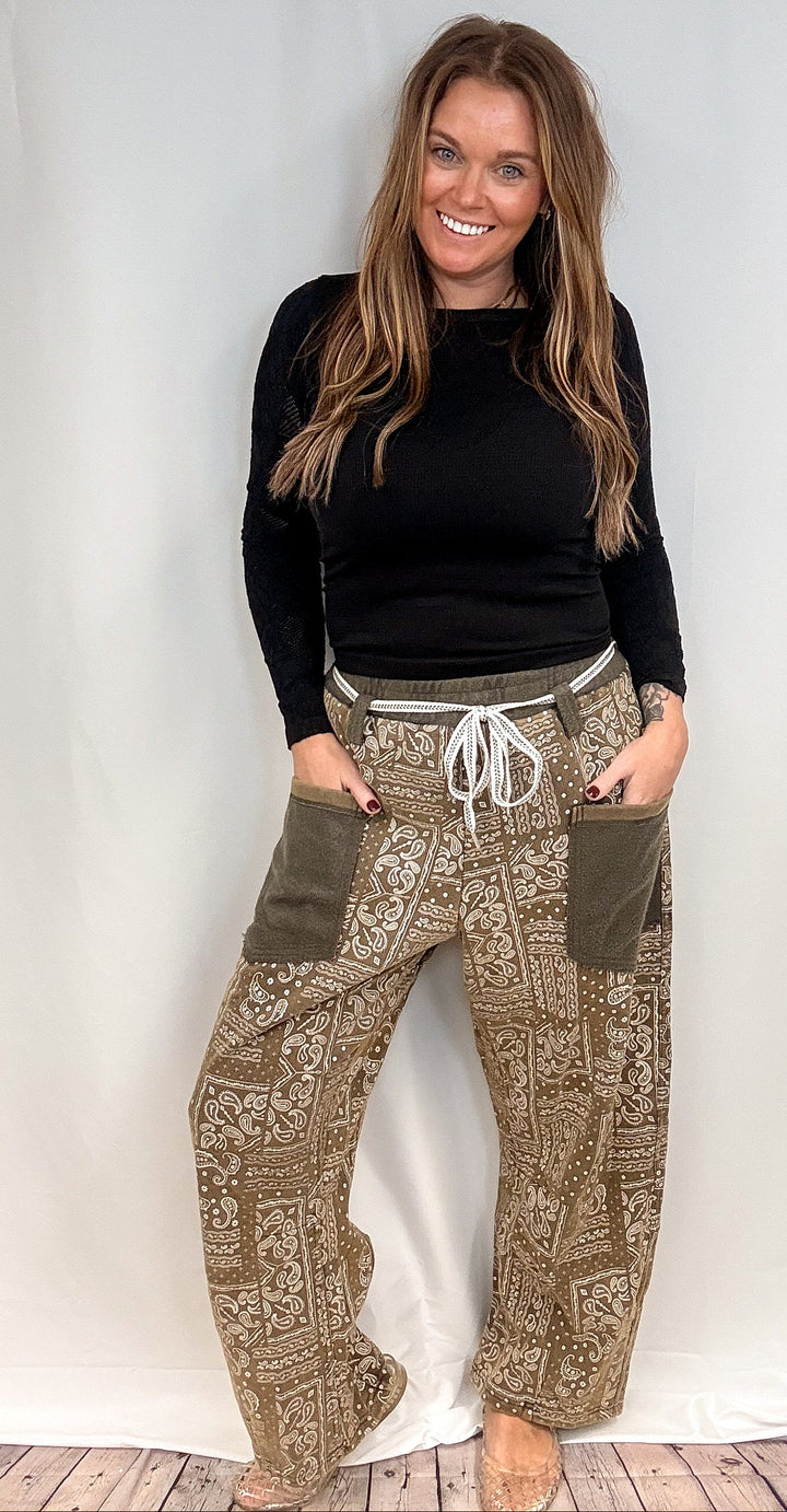 A woman modeling a pair of loose-fit, wide-leg pants with a brown and beige paisley pattern, olive green side pockets, and a white drawstring waist, paired with a black long-sleeve top.