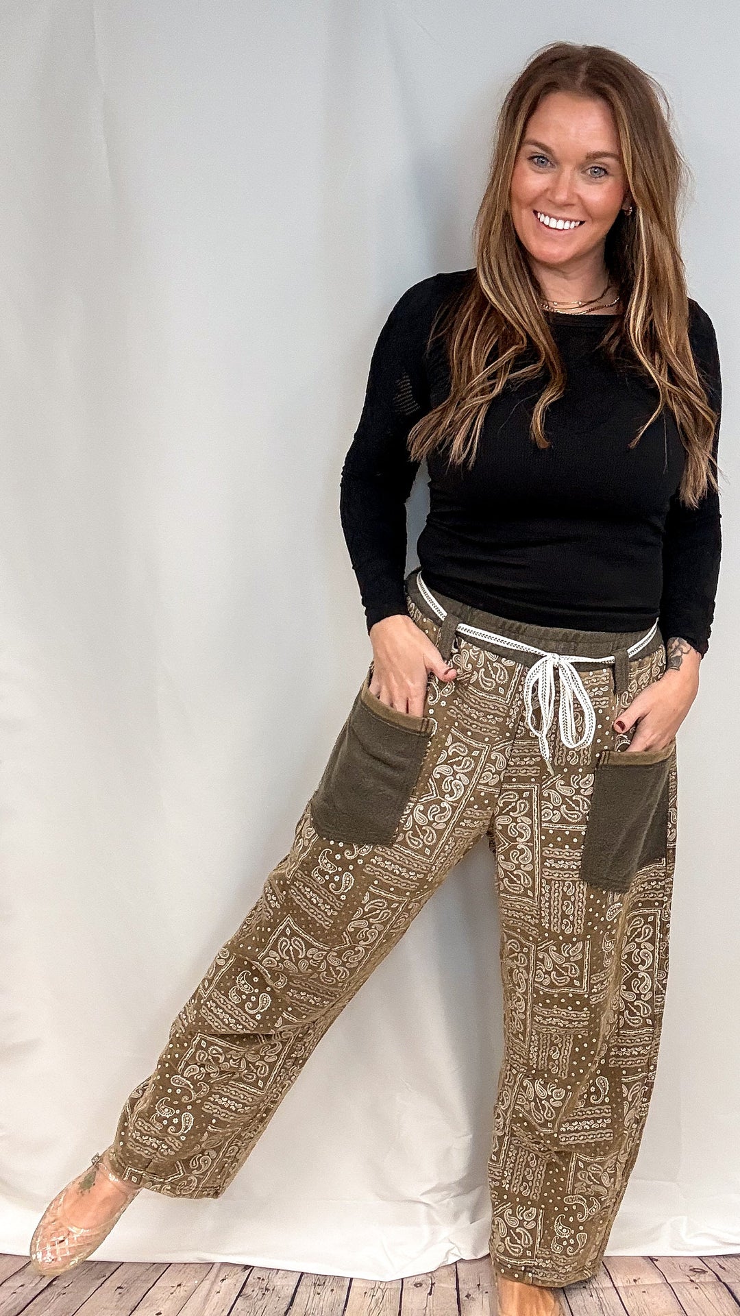 A woman modeling a pair of wide-leg brown pants with a beige paisley pattern, olive green patch pockets, and a white drawstring waist, paired with a black long-sleeve top.