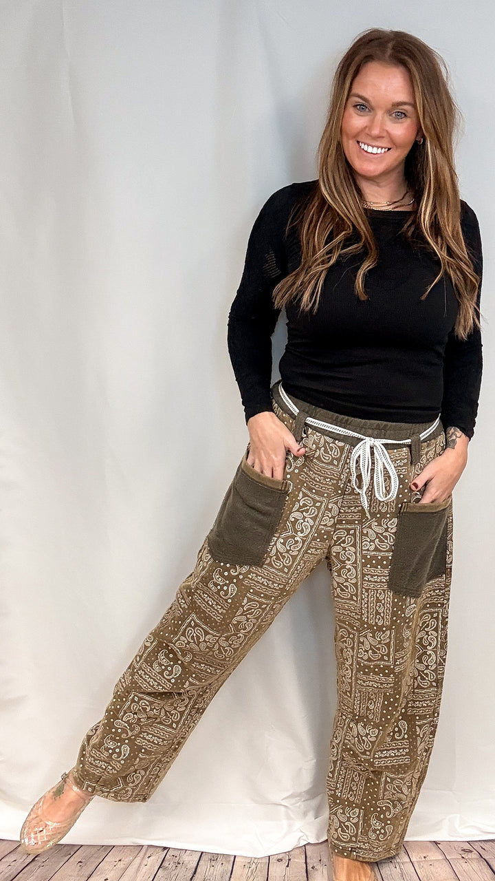 A woman modeling a pair of wide-leg brown pants with a beige paisley pattern, olive green patch pockets, and a white drawstring waist, paired with a black long-sleeve top.