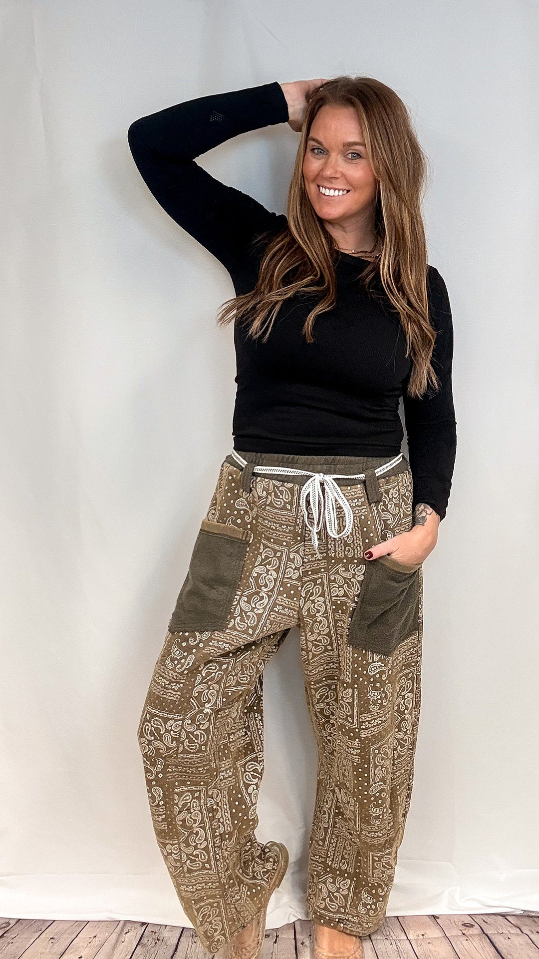 A woman modeling a pair of loose, patterned brown pants with olive green patch pockets and a white drawstring, paired with a black long-sleeve top, posing against a white studio backdrop.