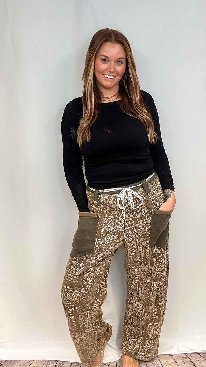 A woman modeling a pair of loose-fit, wide-leg pants with a brown and beige paisley pattern, olive green side pockets, and a white drawstring, paired with a black long-sleeve top against a white studio backdrop.