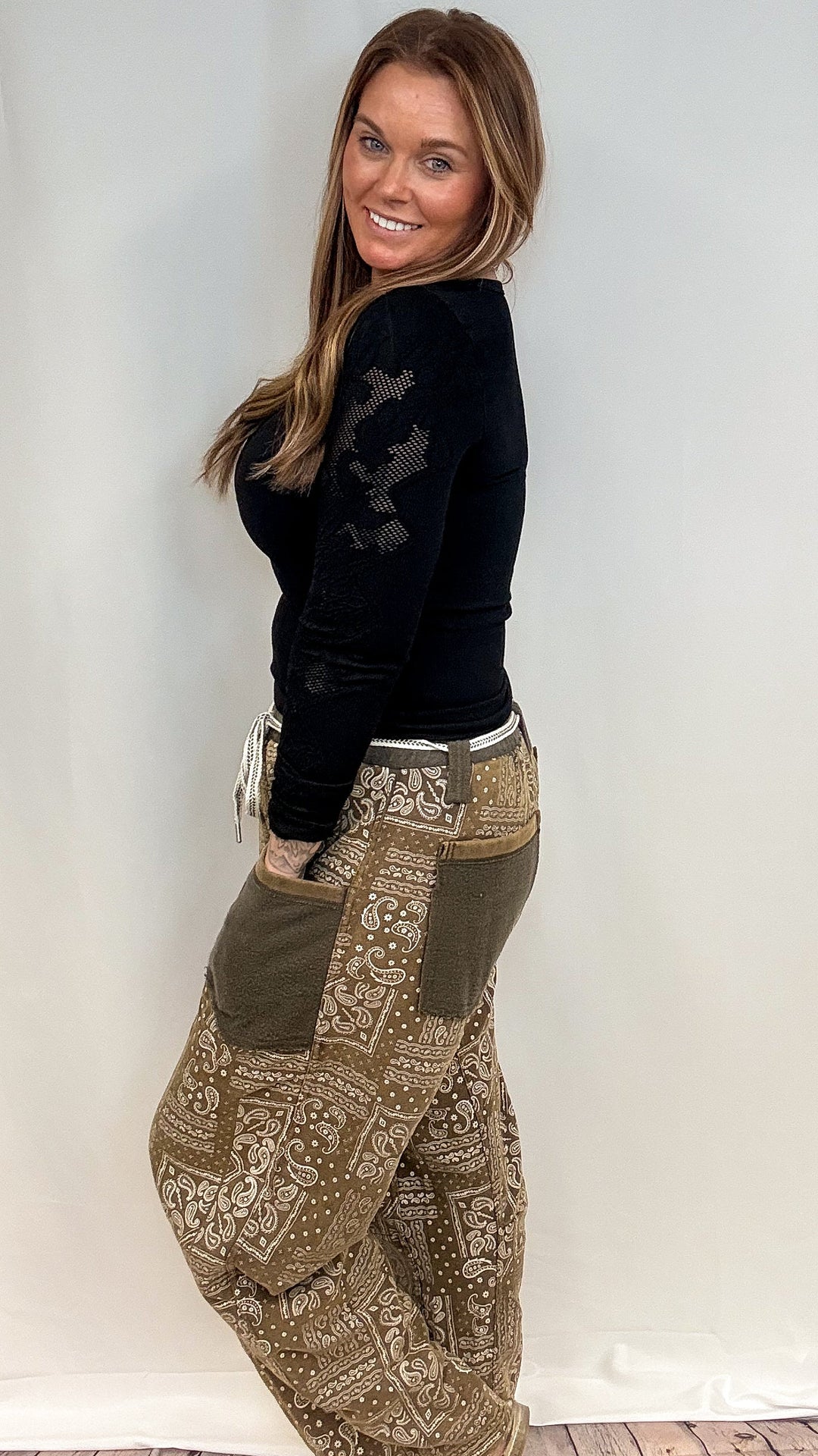 A woman modeling a pair of loose, patterned brown pants with olive green side pockets and a black long-sleeve top, standing against a plain white studio backdrop.