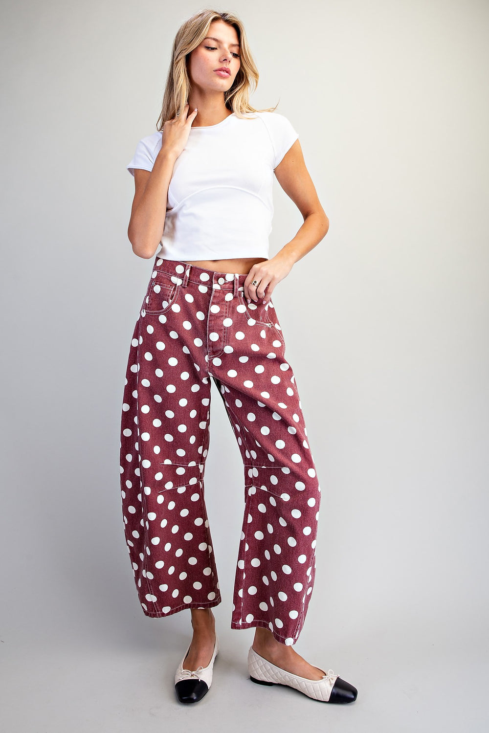 A woman modeling a pair of wide-leg cropped pants in a dusty rose color with white polka dots, paired with a white cropped t-shirt and black-and-white flats, against a light gray studio background.