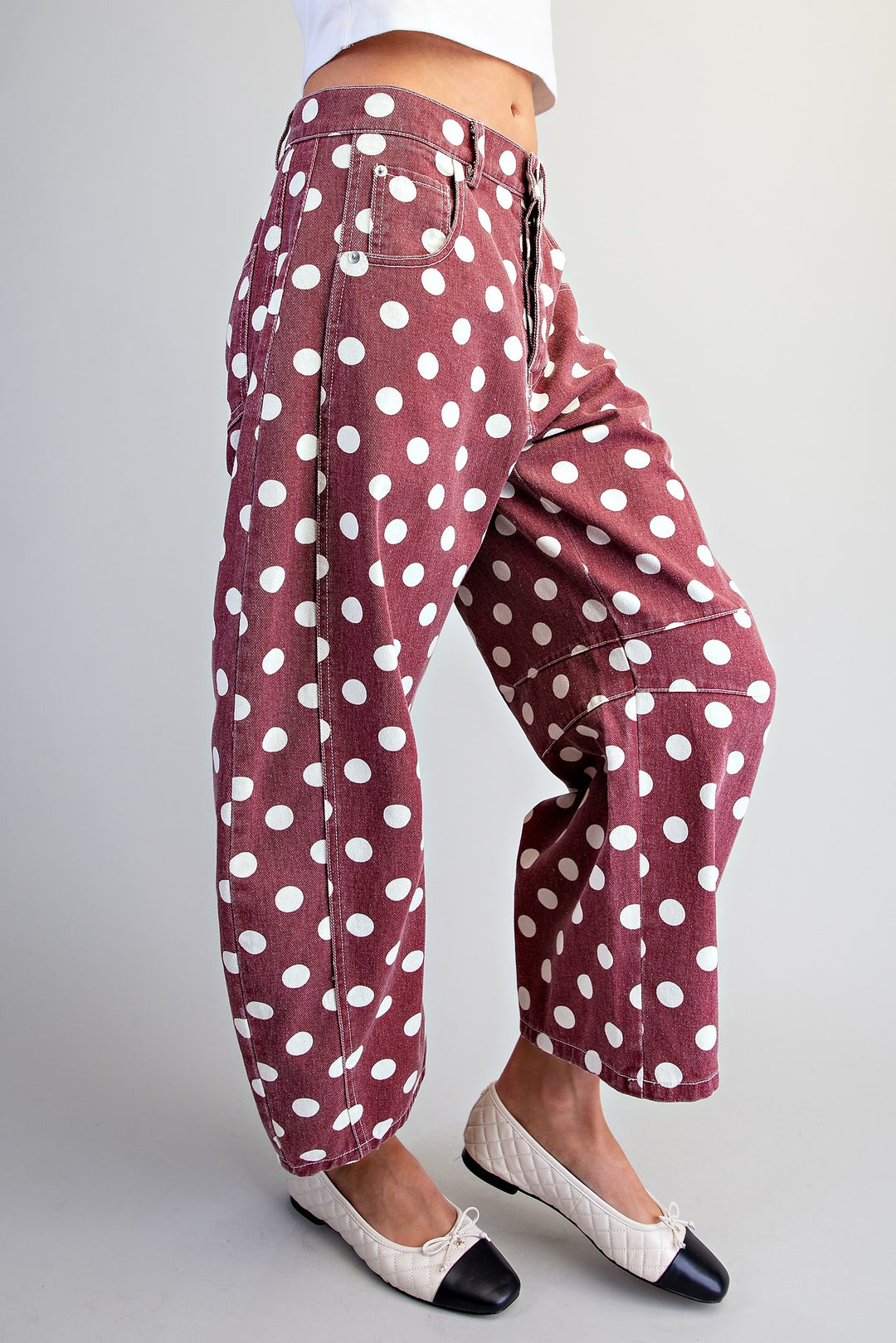 A person wearing loose, wide-leg pants in a dusty rose color with a white polka dot pattern, paired with white and black flat shoes, shown from the waist down against a light gray studio background.