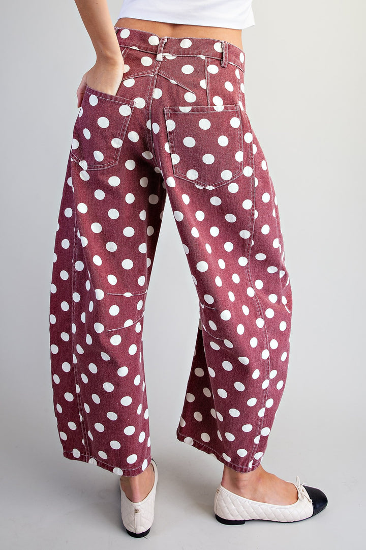 A person wearing loose, wide-leg cropped pants in a dusty rose or mauve color with a white polka dot pattern, paired with white and black flats, shown from the waist down against a light gray studio background.