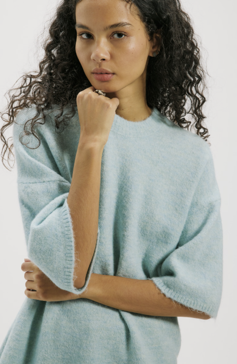 A woman with curly dark hair wearing a light blue fuzzy knit sweater, posing with her hand on her chin against a plain light background.