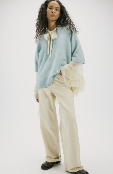 A model stands against a plain light gray studio background wearing a light blue oversized knit sweater with a cream bow at the neckline, layered cream ruffled sheer sleeves, cream wide-leg pants, and black shoes.