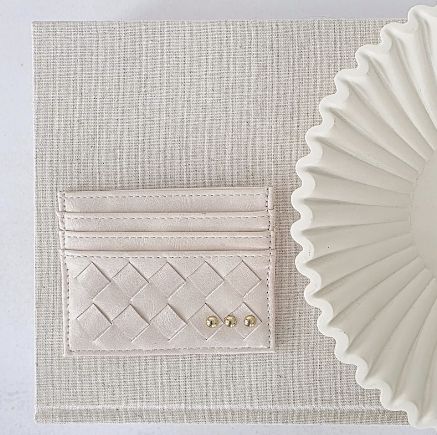 A cream-colored leather card holder with a woven basketweave pattern and two small gold studs, displayed on a textured beige linen background next to a white ceramic plate.