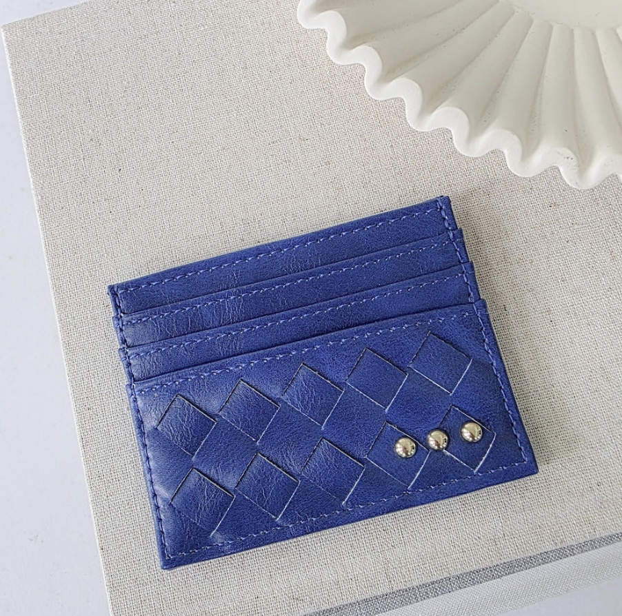 A blue leather card holder wallet with a woven basketweave pattern and two small gold-tone studs, displayed on a textured beige fabric surface.