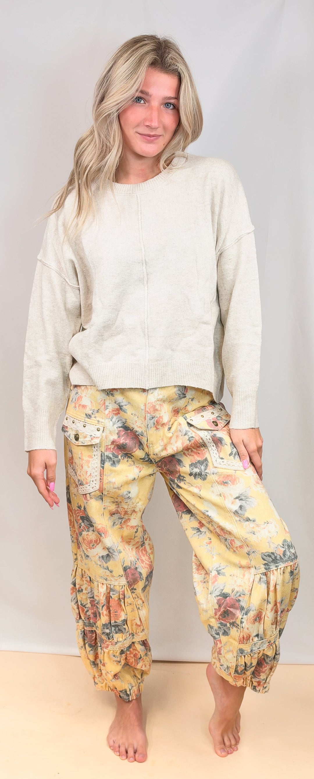 A woman modeling a two-piece loungewear set consisting of a cream-colored long-sleeve top and yellow floral harem pants, standing against a light beige studio backdrop.
