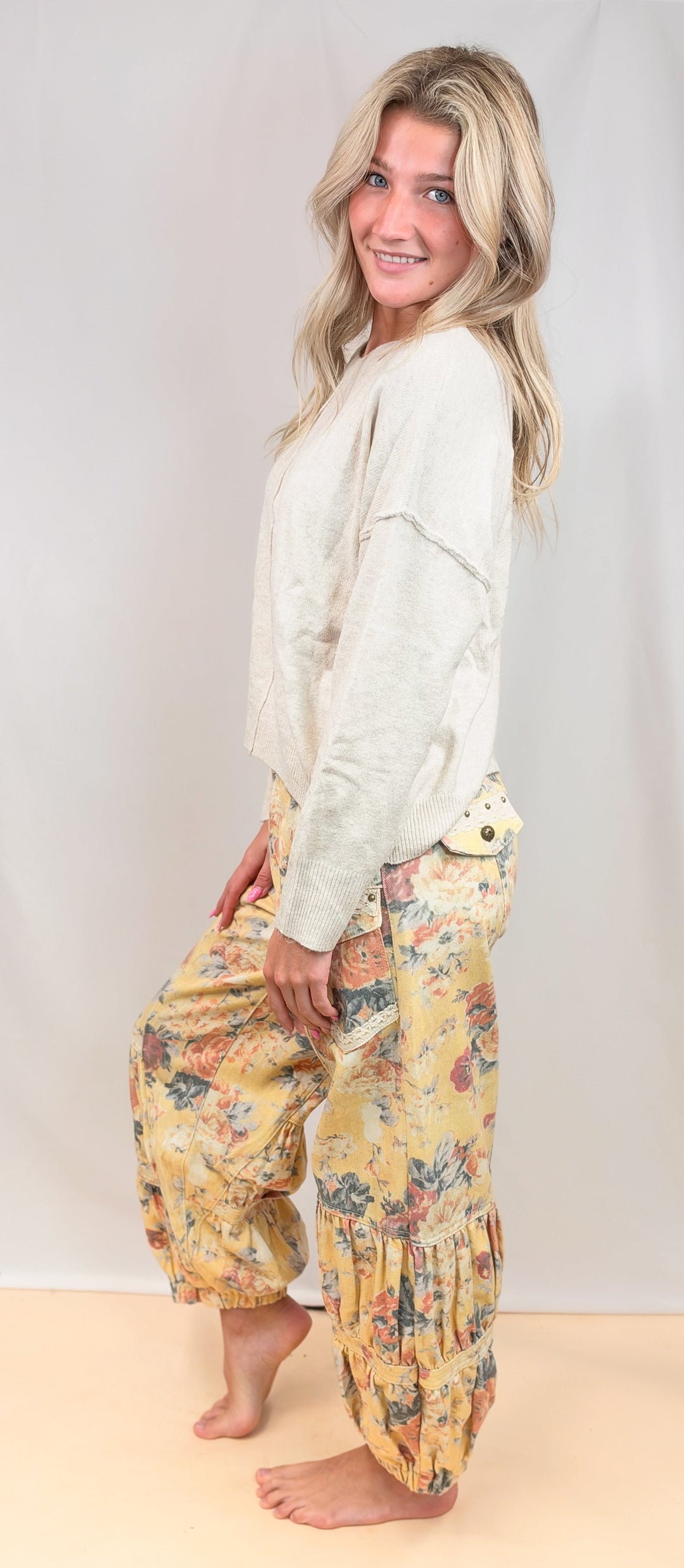 A woman modeling loose, harem-style pants with a yellow and multicolor floral print, paired with a cream-colored long-sleeve top, standing against a light beige studio backdrop.