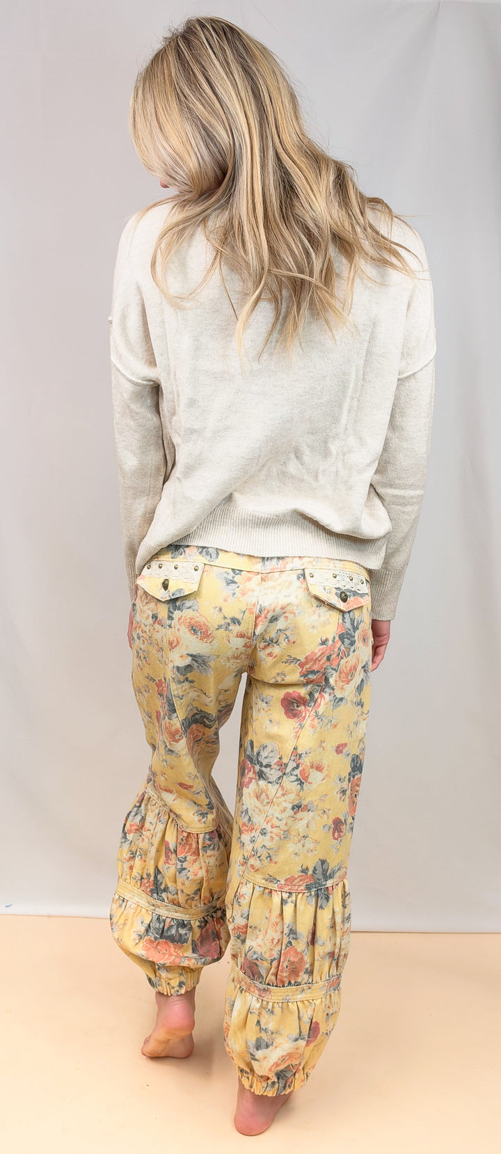 A woman viewed from behind wearing a light beige long-sleeve top and yellow floral print bohemian-style pants with ruffled hems, standing barefoot against a neutral studio backdrop.