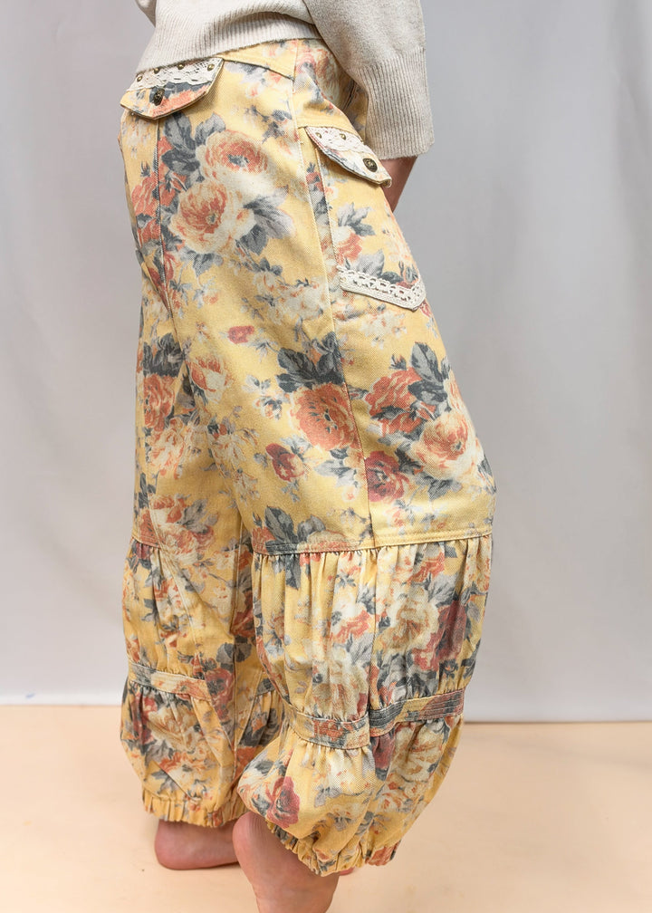 A person wearing loose, balloon-style floral pants with elastic cuffs and decorative lace trim, standing barefoot against a plain studio backdrop.