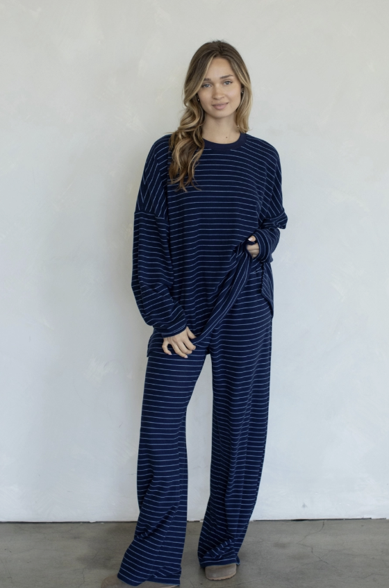 A woman modeling a matching navy blue and white striped lounge set, consisting of a long-sleeve top and wide-leg pants, standing against a textured white wall.