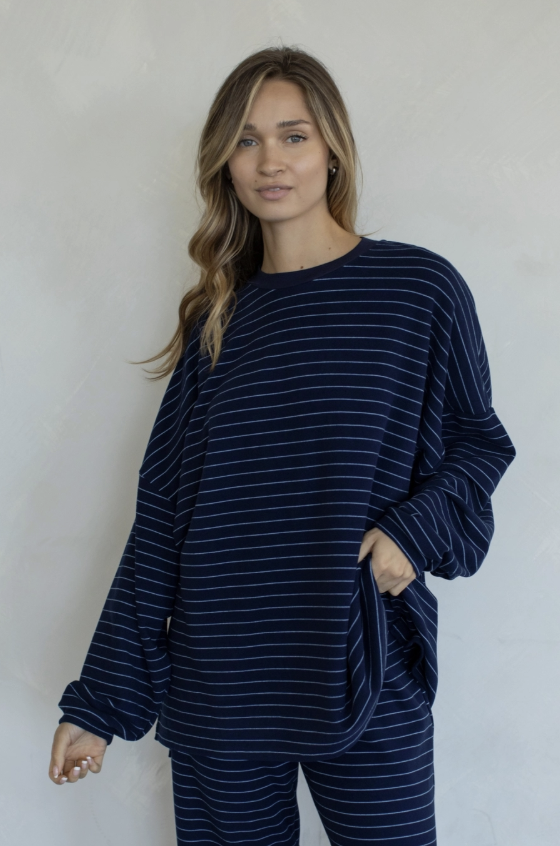 A woman modeling a matching navy blue and white striped lounge set, featuring a long-sleeve top and pants, against a textured off-white wall.