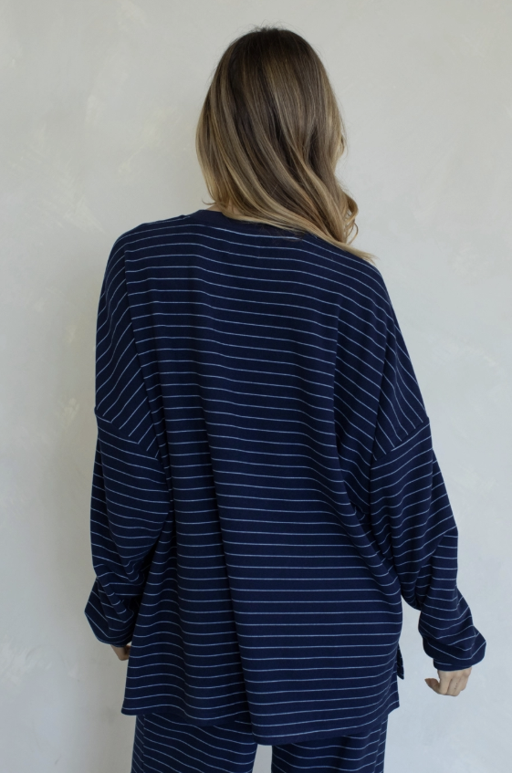 A woman viewed from behind wearing a navy blue long-sleeve top with thin white horizontal stripes and matching striped pants against a light textured wall.