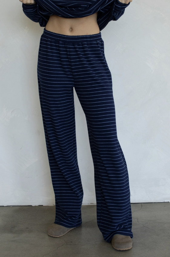 A person wearing loose navy blue pants with thin white horizontal stripes, paired with a matching navy top and tan slippers, standing against a white wall.