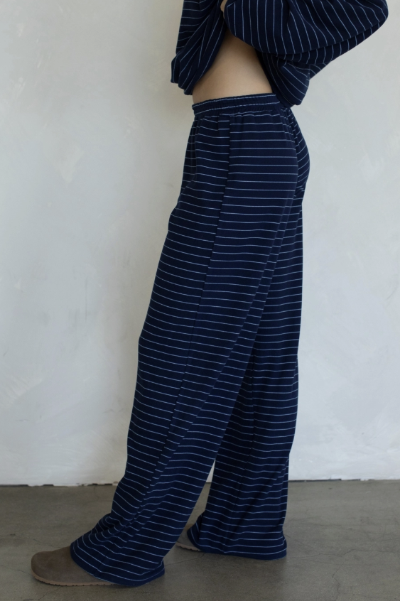 A person wearing loose, wide-leg pants with thin white horizontal stripes on a dark navy blue fabric, paired with a matching navy top and brown slippers, standing against a textured off-white wall.