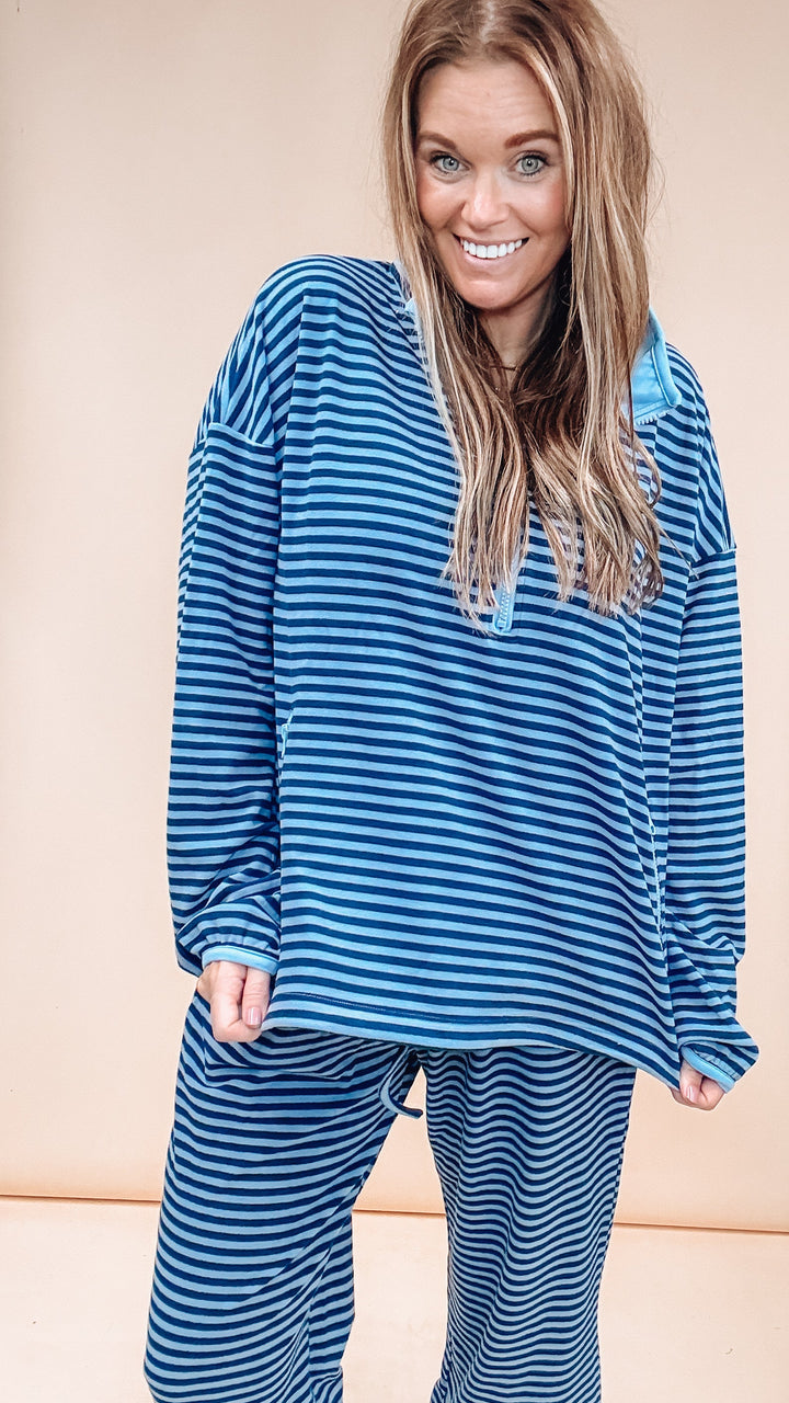 A woman modeling a matching two-piece pajama set with blue and navy horizontal stripes, featuring a long-sleeve top and pants, against a solid beige background.