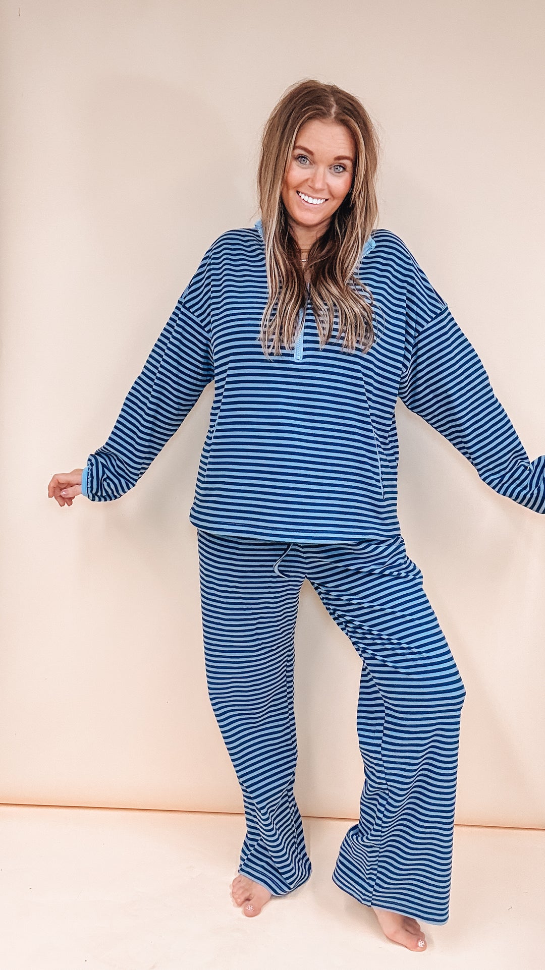 A woman modeling a matching two-piece pajama set with blue and white horizontal stripes, consisting of a long-sleeve top and matching pants, against a plain beige background.