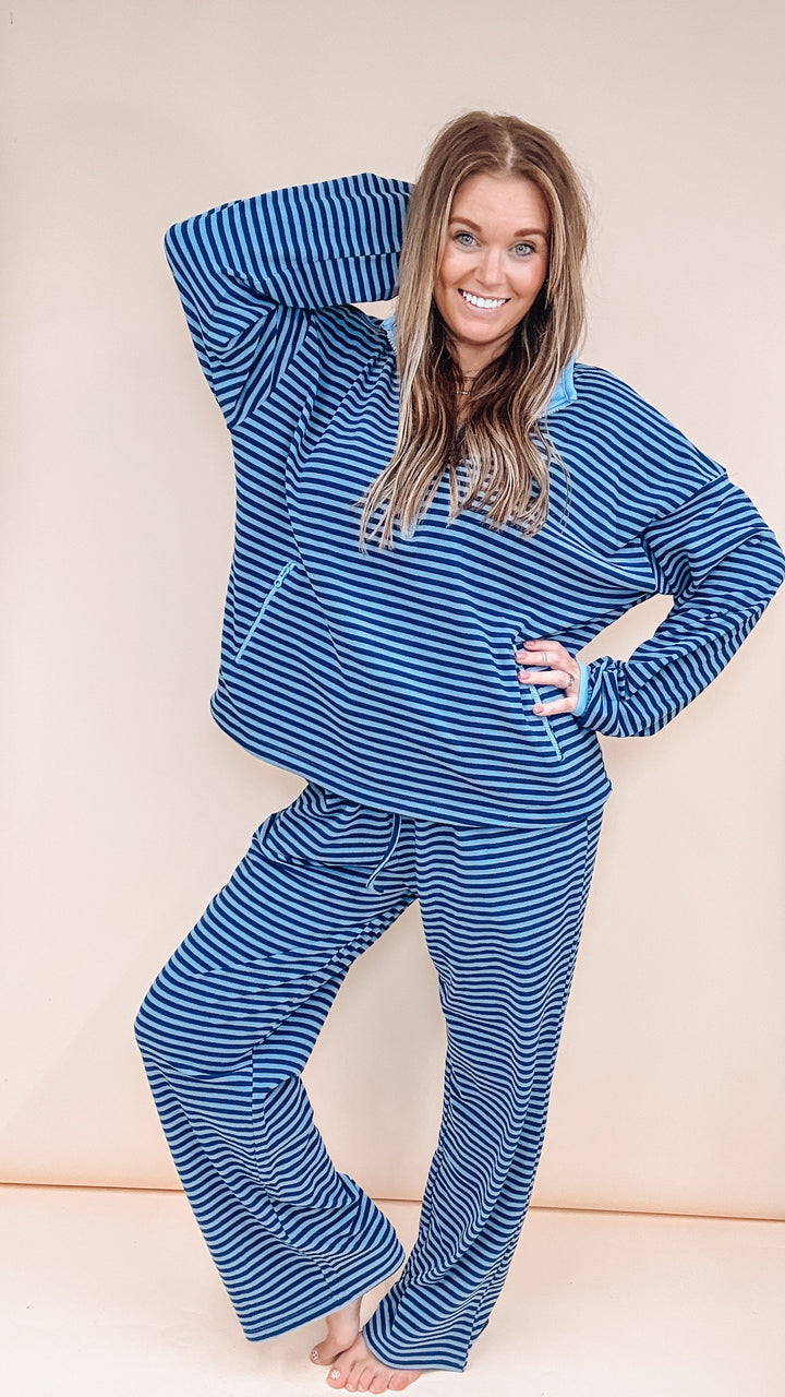 A woman modeling a matching two-piece pajama set with blue and white horizontal stripes, featuring a long-sleeve top and wide-leg pants, posed against a solid light beige background.