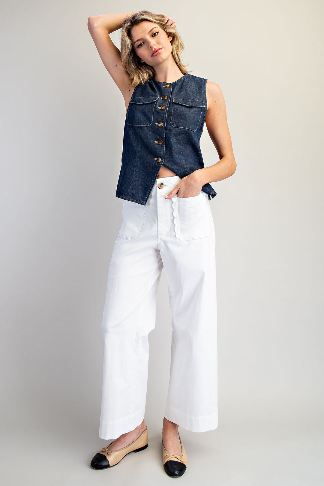 Woman wearing a denim vest over a white top with white pants on a plain background