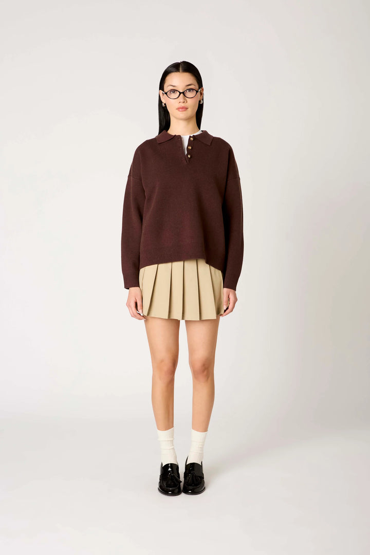 A model stands against a light gray studio background wearing a dark brown mock-neck knit sweater, a pleated beige mini skirt, white socks, and black loafers.