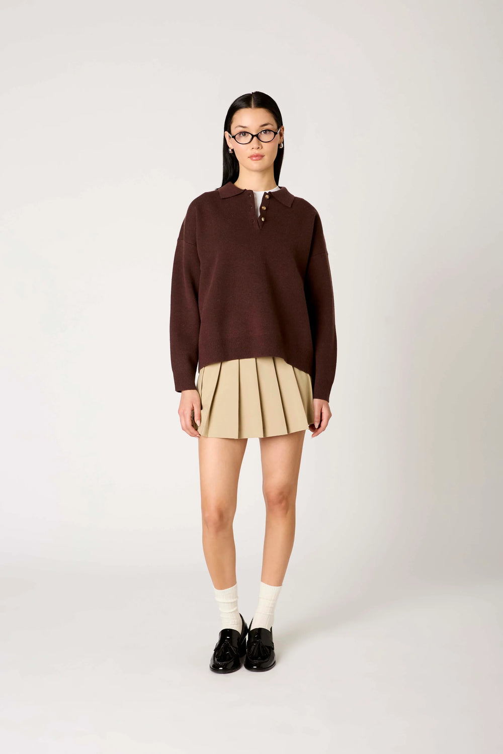 A model stands against a light gray studio background wearing a dark brown long-sleeve mock-neck sweater, a beige pleated mini skirt, white socks, and black loafers.