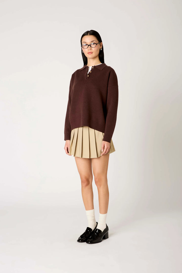 A model wearing a dark brown mock-neck knit sweater, a beige pleated mini skirt, white socks, and black loafers, standing against a plain light gray studio background.