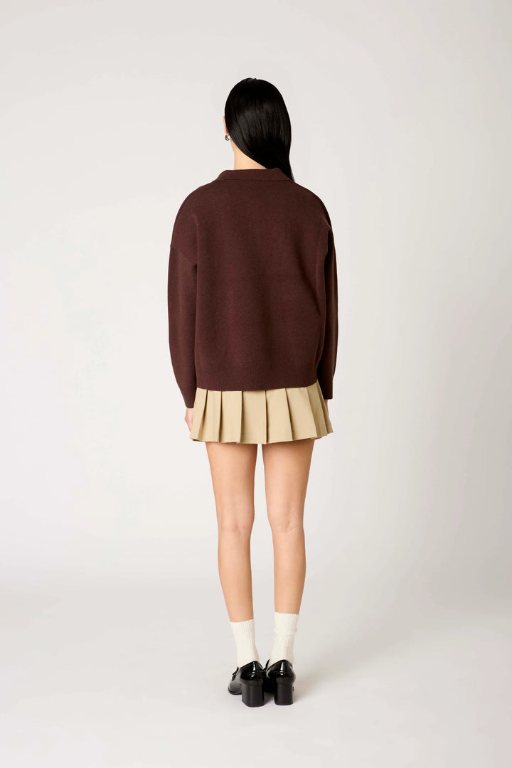 A model shown from the back wearing a dark brown knit sweater, a pleated beige mini skirt, white socks, and black shoes against a light gray studio background.