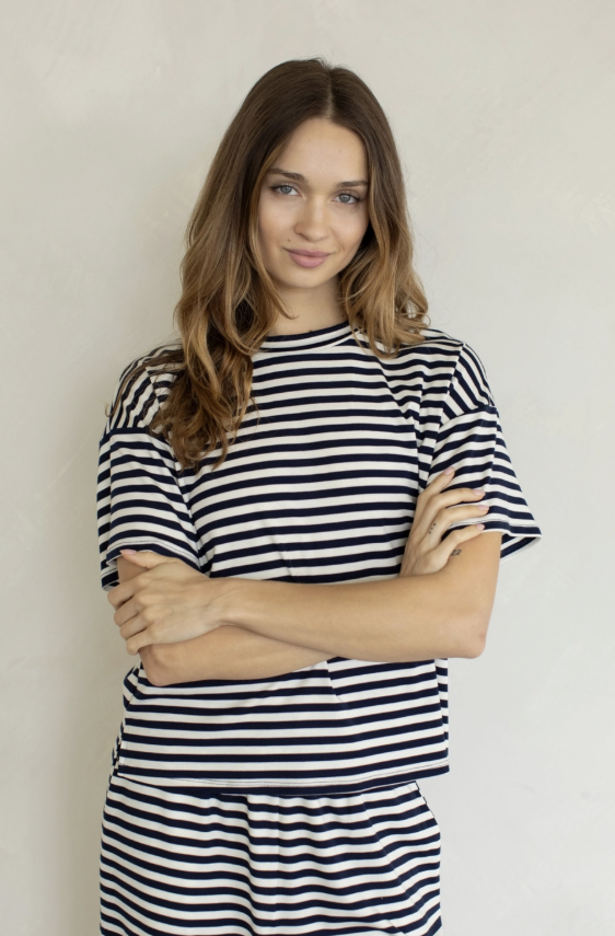 A woman with long brown hair wearing a black and white striped short-sleeve top and matching bottoms, standing with her arms crossed against a light textured wall.