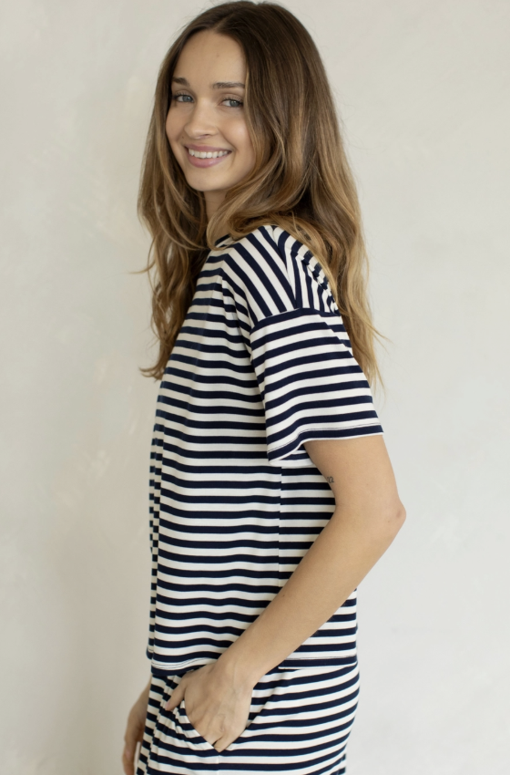 A woman with long light brown hair is shown from the side and back, wearing a short-sleeve navy and white horizontally striped t-shirt dress against a plain off-white wall.