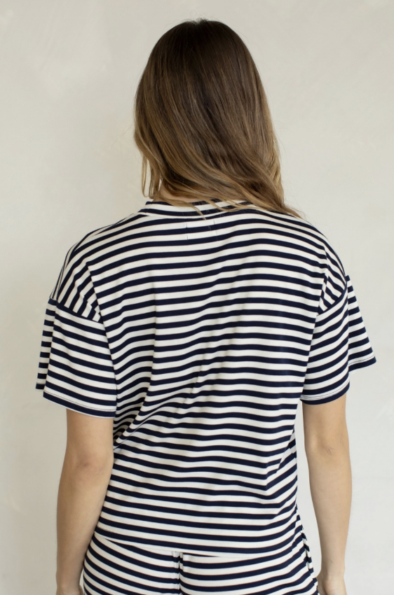 A woman viewed from behind wearing a short-sleeve striped T-shirt with horizontal navy and off-white stripes, set against a plain light beige wall.