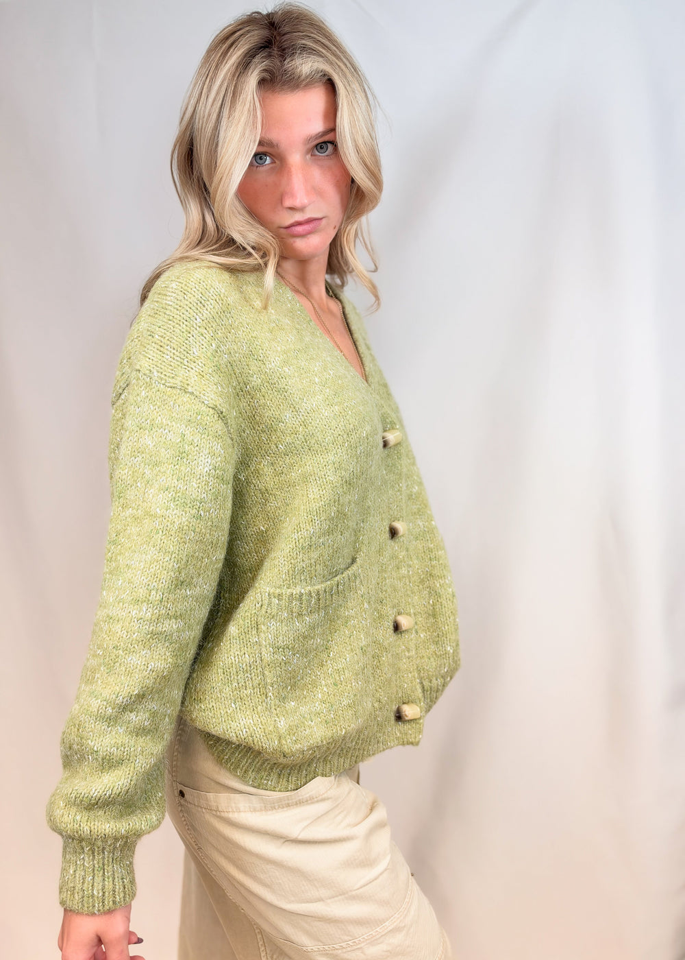 Woman shown from side view wearing green cardigan sweater with beige pants on plain background