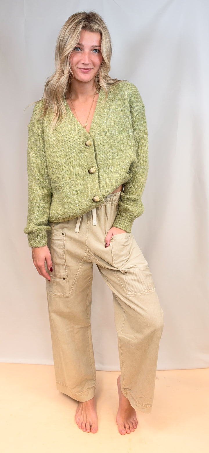 Woman wearing green cardigan sweater with large buttons and beige pants on plain background
