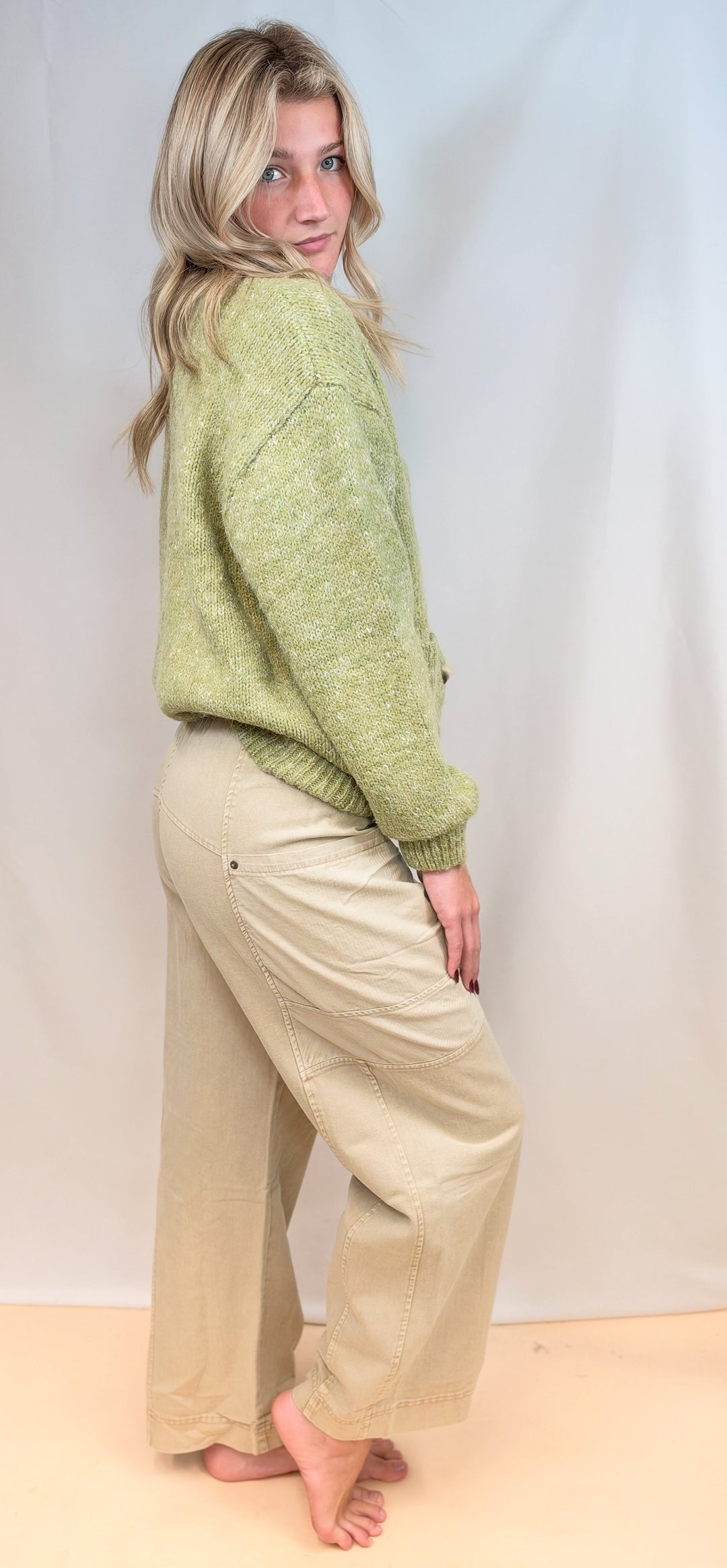 Woman shown from side view wearing green cardigan sweater and beige pants on plain background