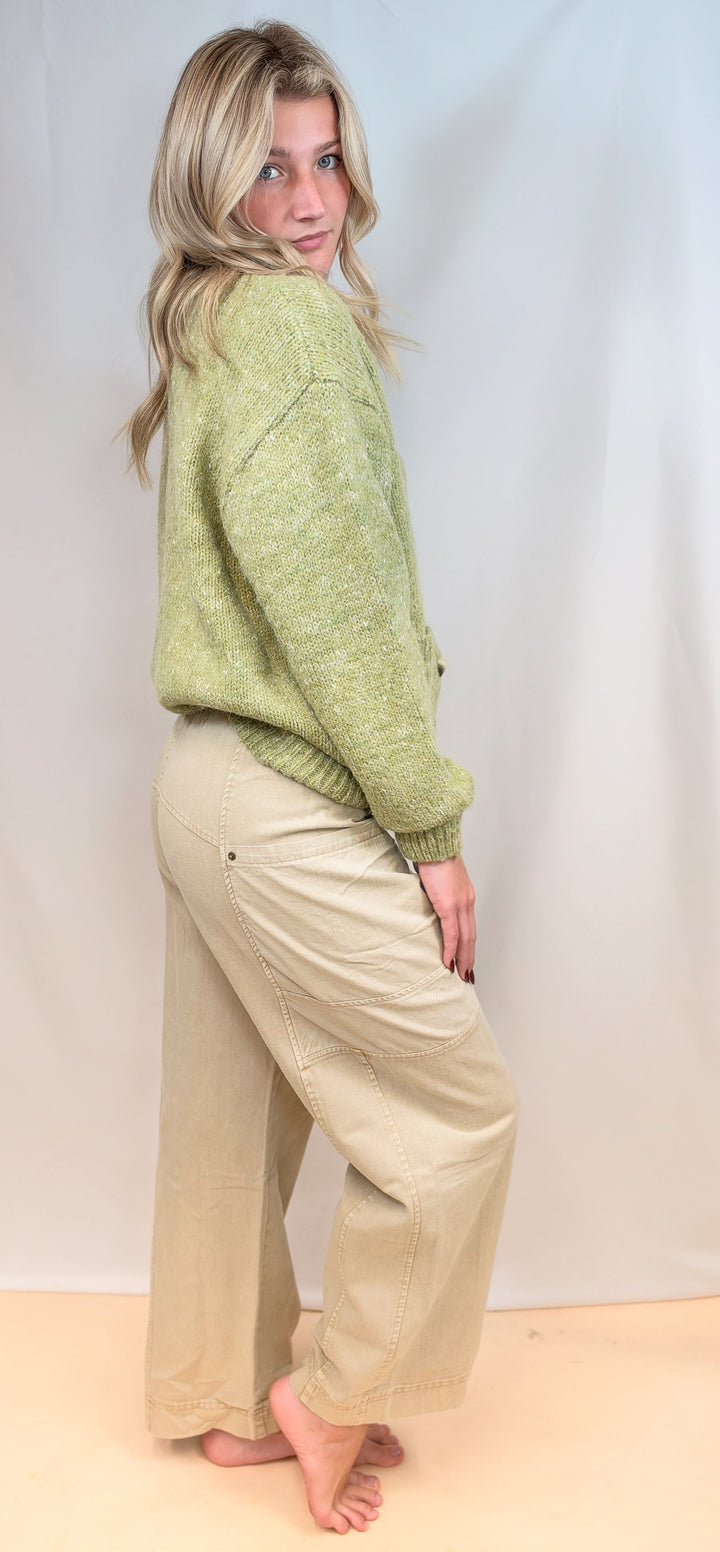 Woman shown from side view wearing green cardigan sweater and beige pants on plain background