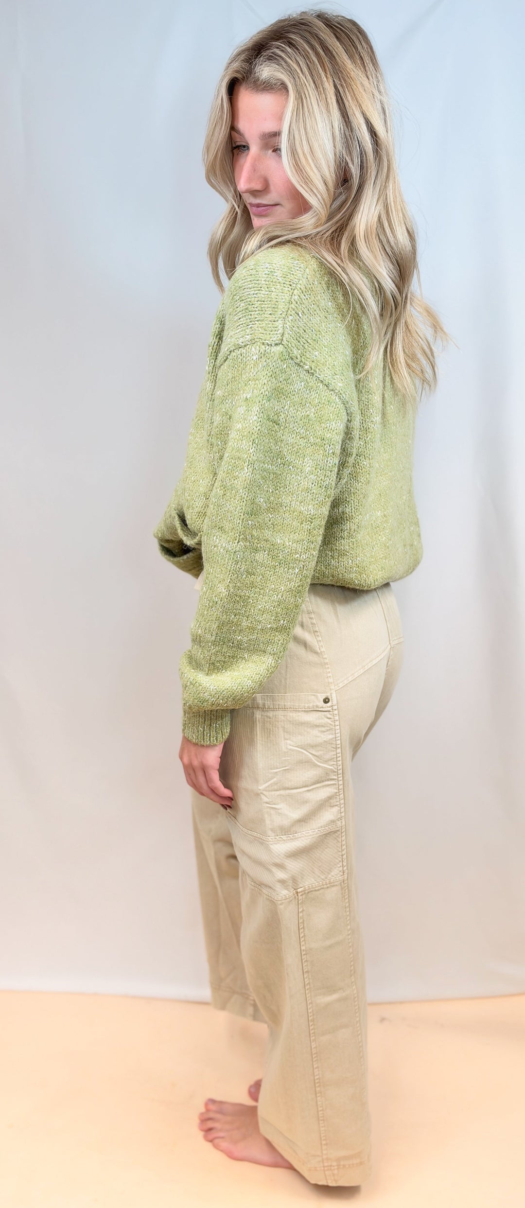 Woman shown from side view wearing green cardigan sweater and beige pants on plain background