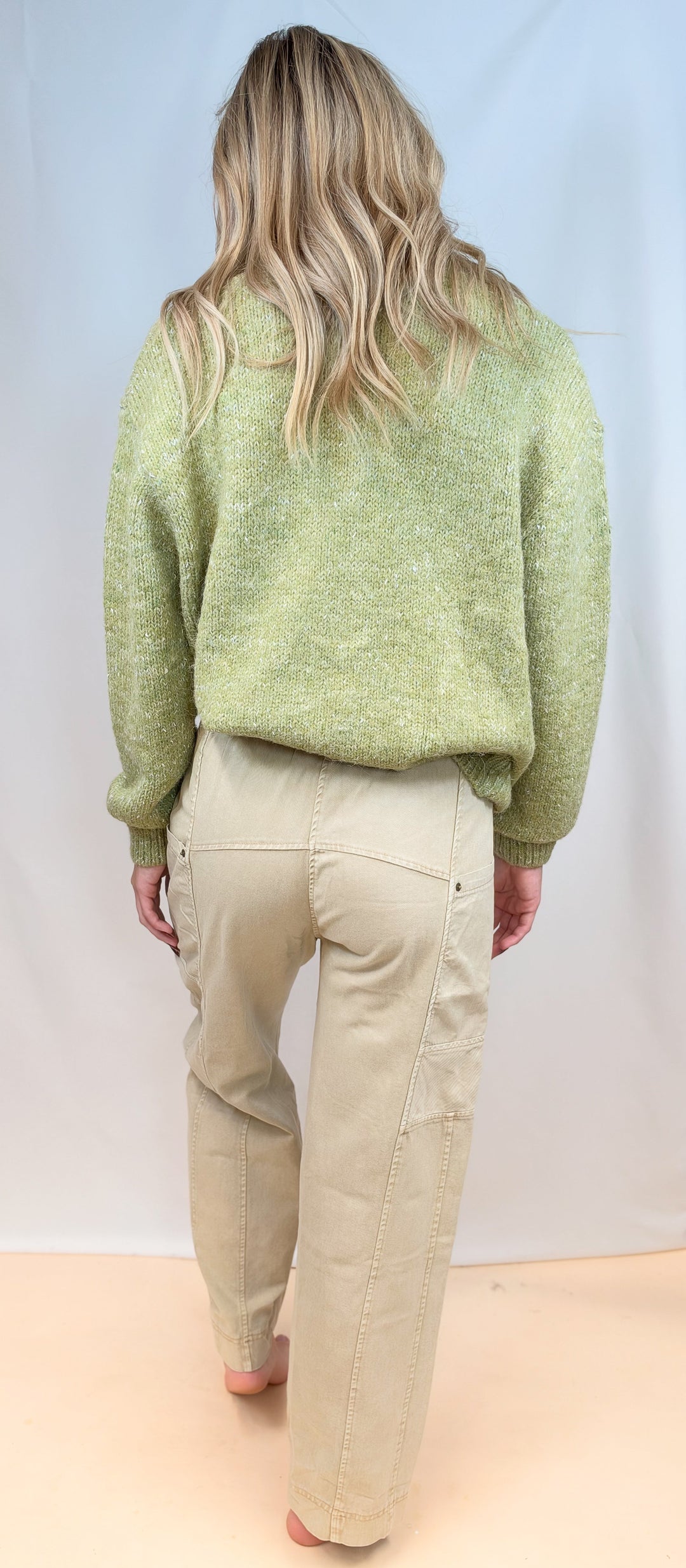 Woman shown from back view wearing green cardigan sweater with beige pants on plain background