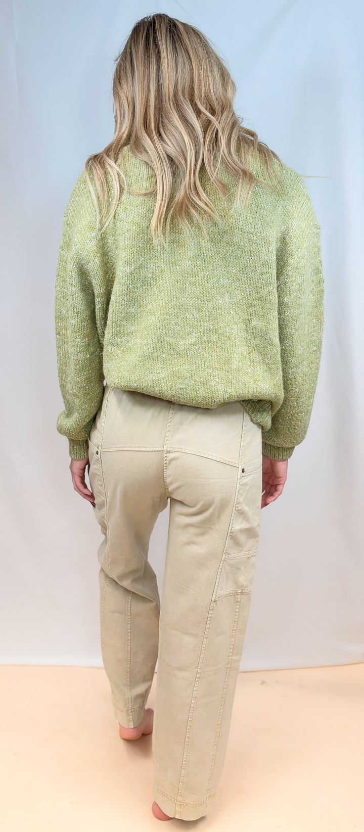 Woman shown from back view wearing green cardigan sweater with beige pants on plain background