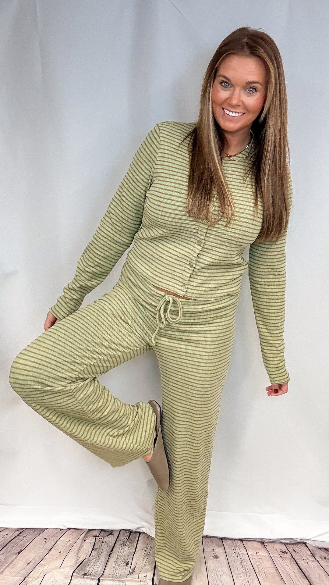 A woman modeling a matching two-piece lounge set with a long-sleeve top and drawstring pants in a muted sage green and cream stripe pattern, posed against a white studio backdrop.