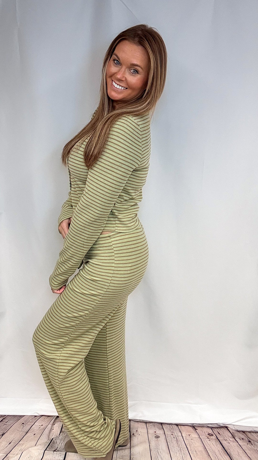 A woman modeling a matching two-piece lounge set with a long-sleeve top and wide-leg pants, featuring a muted olive green and beige horizontal stripe pattern, posed against a white studio backdrop.