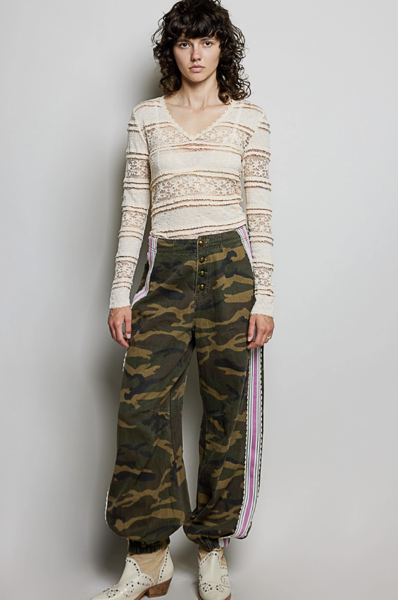 Woman wearing camo pants with pink ribbon detail and beige top on plain background