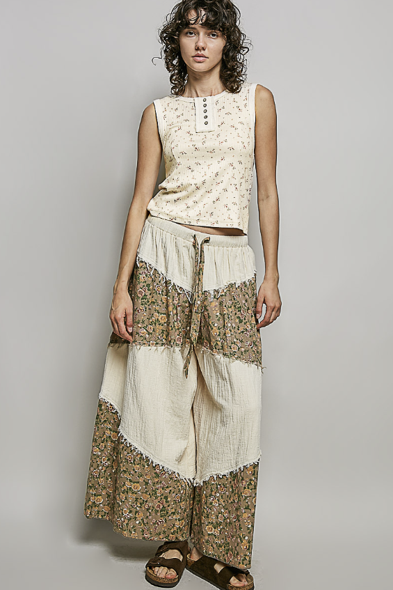 A woman modeling a sleeveless cream top with a small floral print and matching wide-leg cropped pants with beige and brown floral border panels, photographed against a plain light gray studio background.