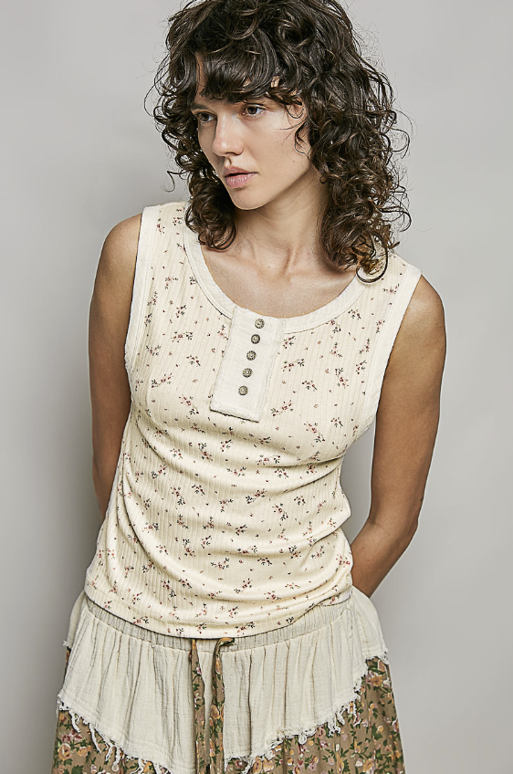 A woman wearing a sleeveless cream-colored top with a small floral print and a buttoned henley-style neckline, paired with a fringed skirt, against a plain gray studio background.