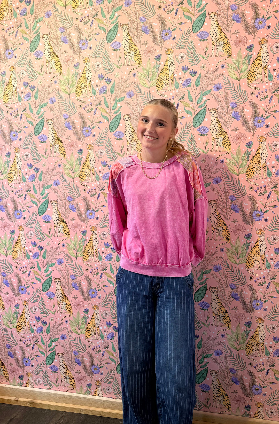 A woman wearing a pink long-sleeve sweatshirt and blue jeans stands in front of a pink leopard-print wallpaper background.