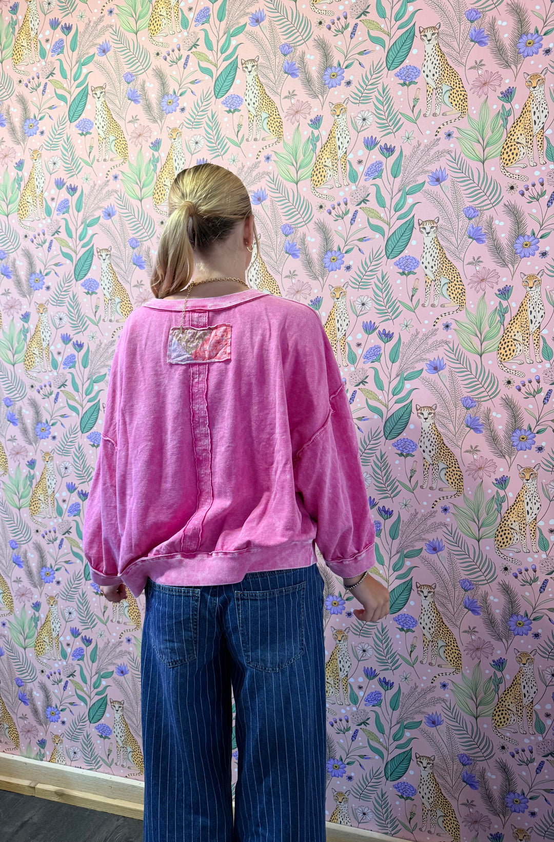 A woman viewed from behind wearing a pink long-sleeve button-up shirt with a decorative patch on the back and blue pinstriped trousers, standing in front of a pink wallpapered wall with leopard prints.
