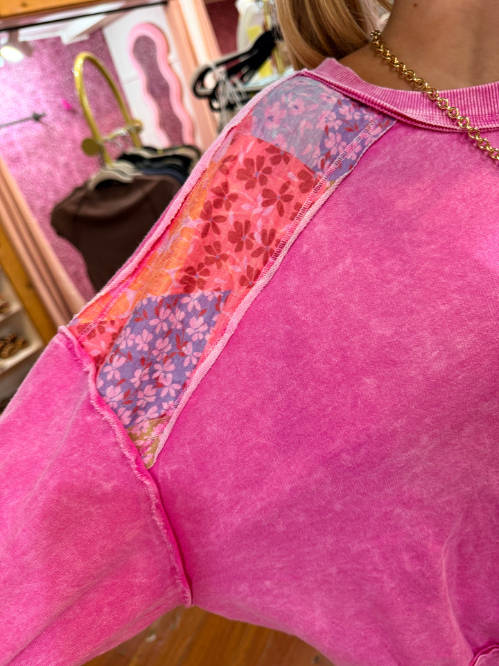A close-up of a pink textured top or dress featuring a decorative patchwork fabric panel with floral and paisley patterns in pink, orange, and purple, worn by a person.