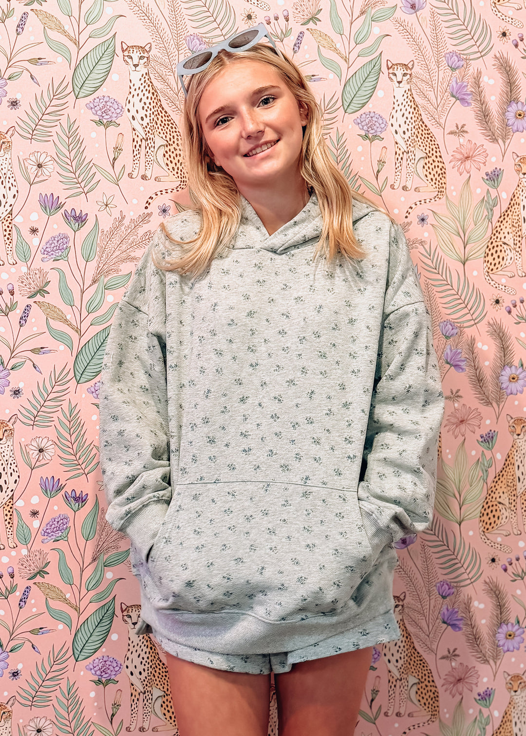 A young woman with blonde hair and sunglasses on her head stands in front of a pink floral and cheetah-patterned backdrop, wearing a heather grey patterned hoodie and matching shorts.
